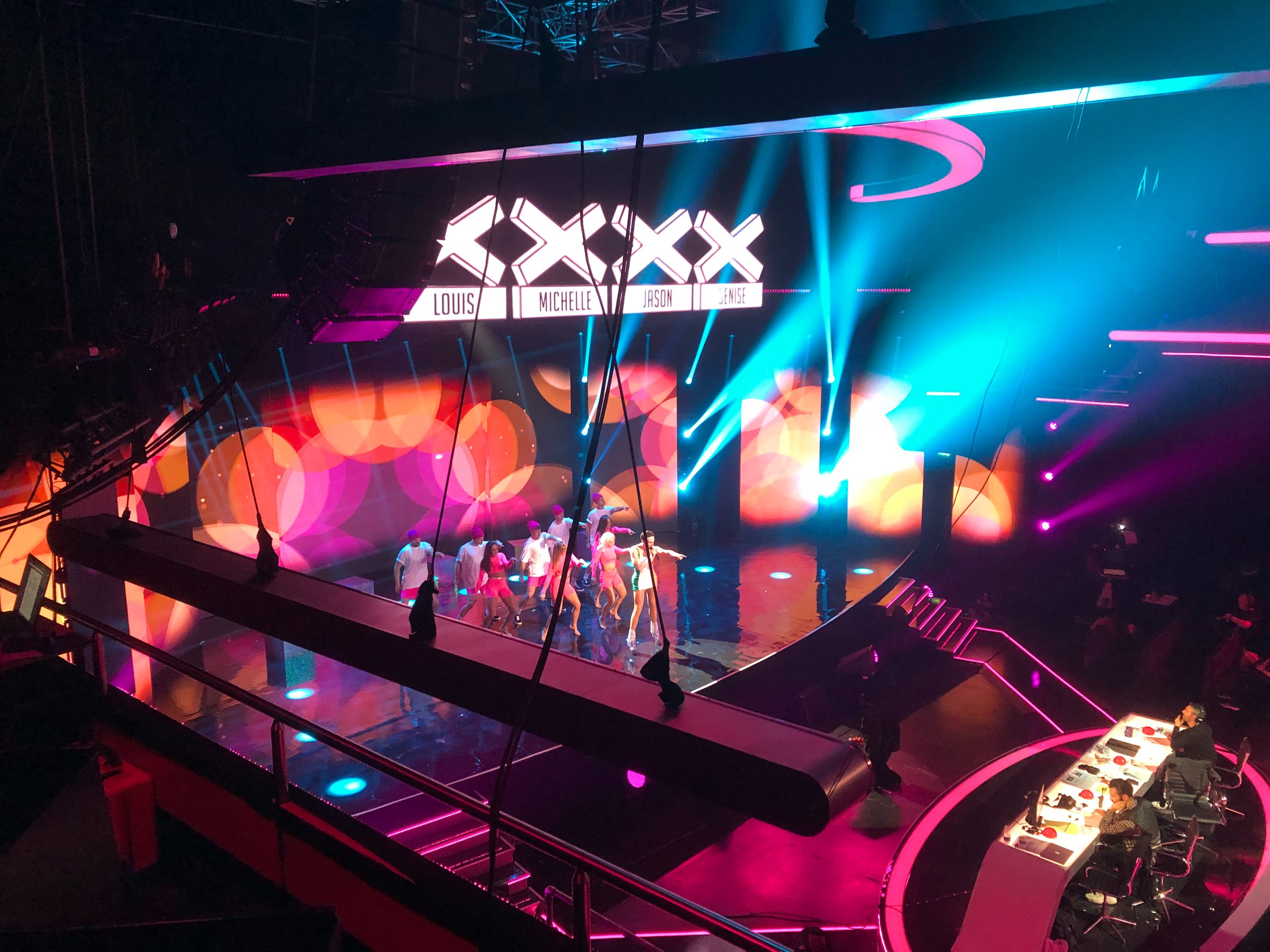 A performer ramp lit in pink and blue beneath a large video screen on a television entertainment set, with a judges' desk in the foreground.