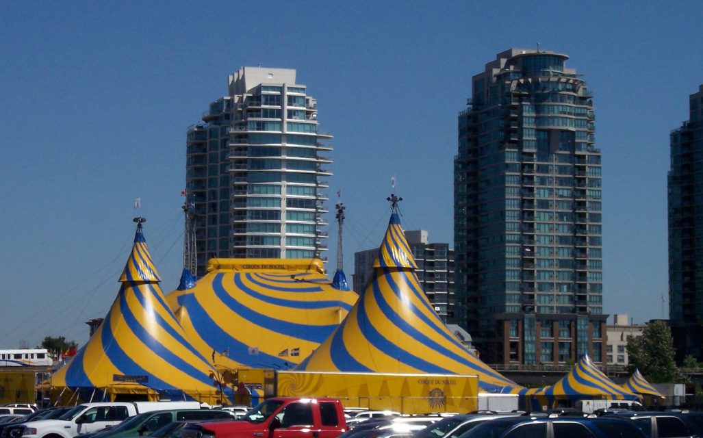 A Cirque du Soleil big top tent by day, with city tower blocks behind.