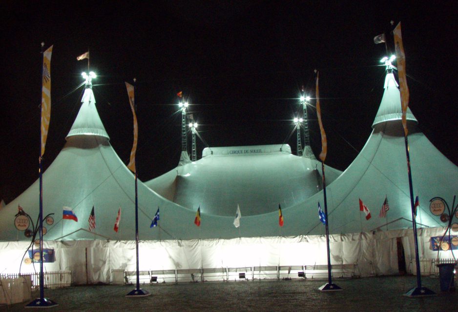 A Cirque du Soleil big top tent lit up at night, flanked by national flags.