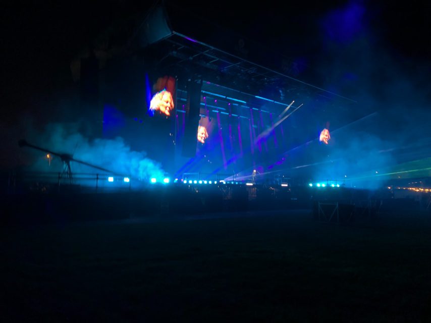 A festival stage at night with a performer's face shown on tall video screens through haze and lighting.