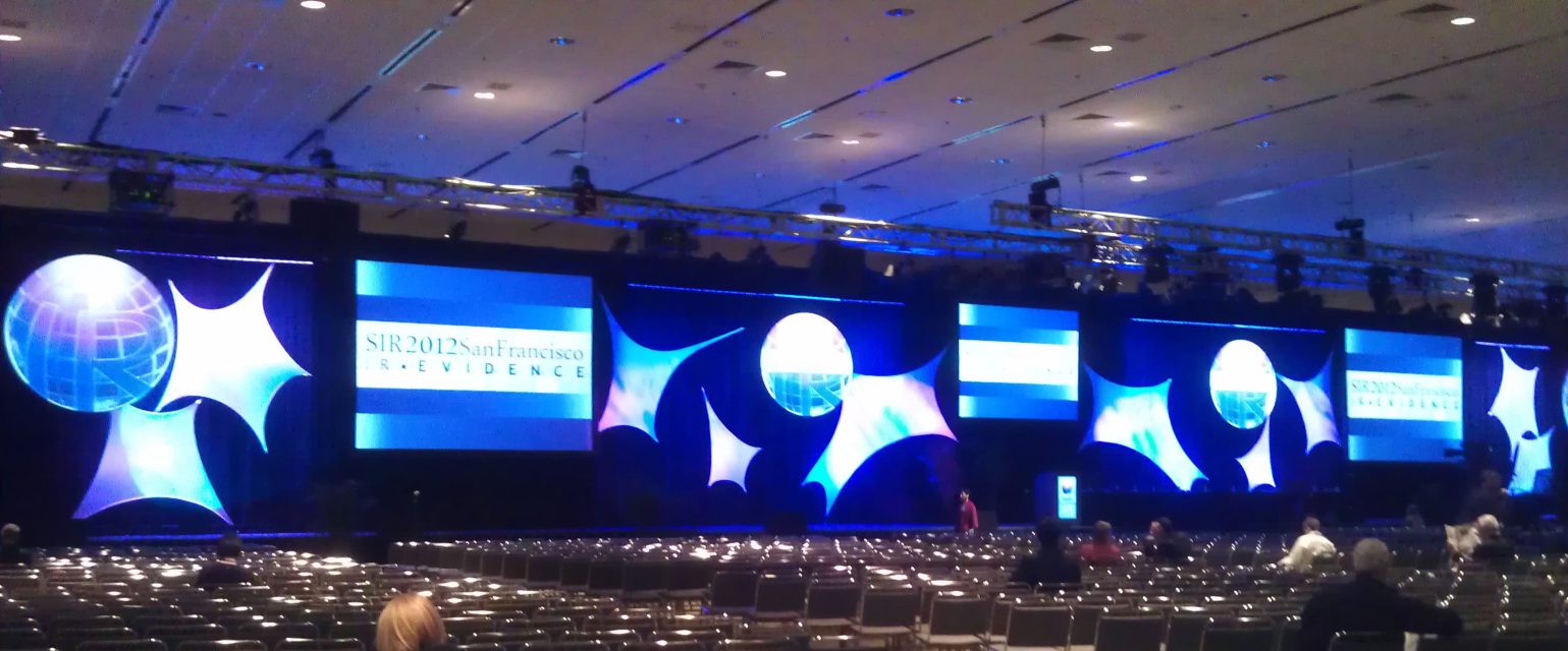 A conference stage with a row of large video screens above an audience at a corporate general session.