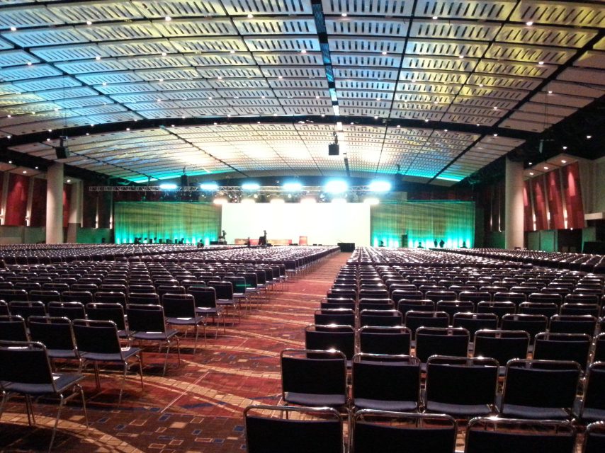 A large empty conference hall set with rows of chairs facing a lit stage.