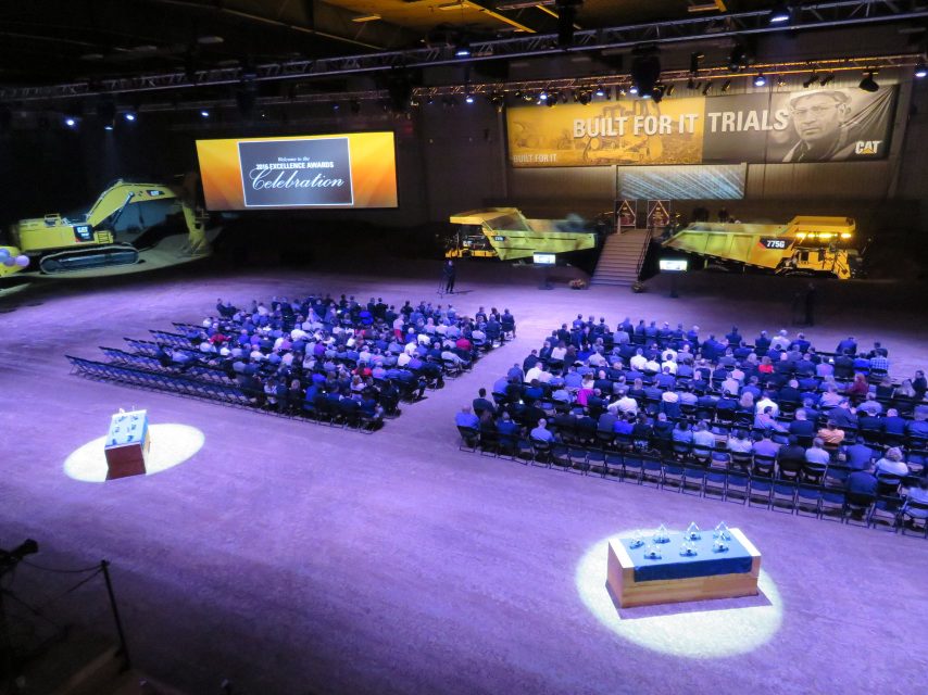 An audience seated before a stage and screens at a corporate event, with heavy equipment displayed either side.