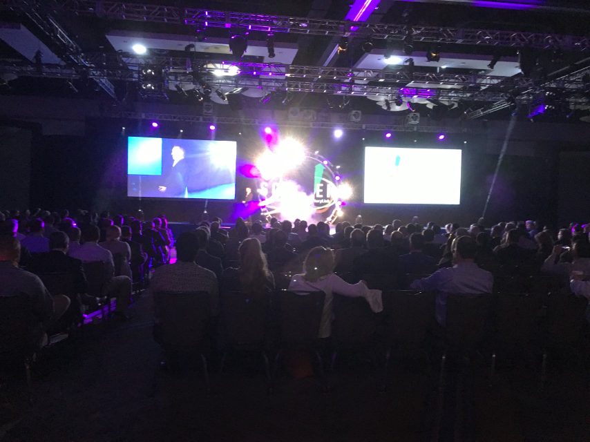 A corporate conference stage with a bright pyrotechnic-style light burst above an audience.