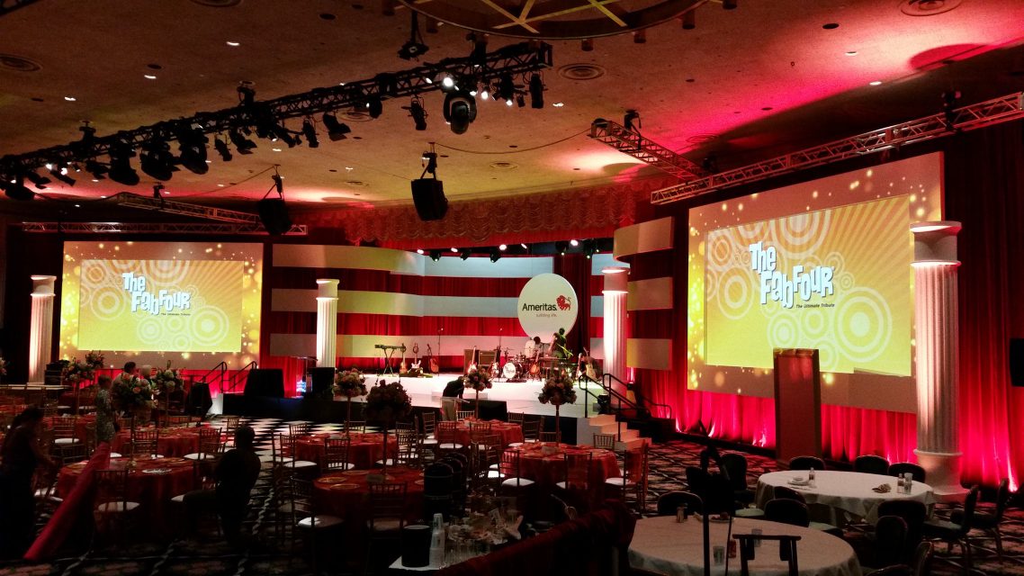 A hotel ballroom set for a banquet, with video screens reading 'The Fab Four' either side of a band stage.