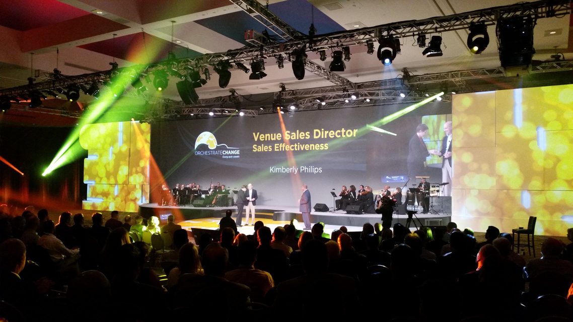 An awards-style stage with a live band and video screens reading 'Venue Sales Director'.