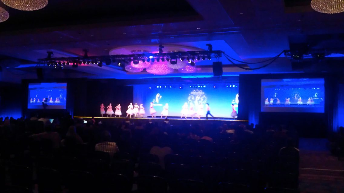 A theatre stage lit in blue with a line of dancers performing beneath video screens.