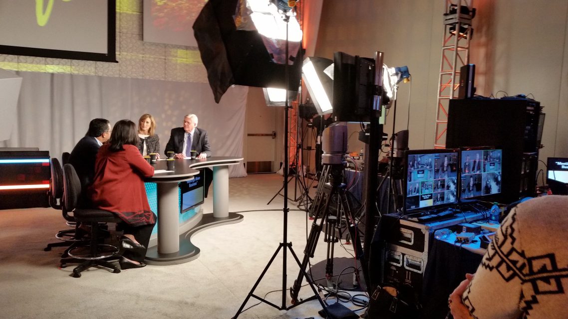 A television studio set with a panel of four seated guests, cameras, and lighting in the foreground.