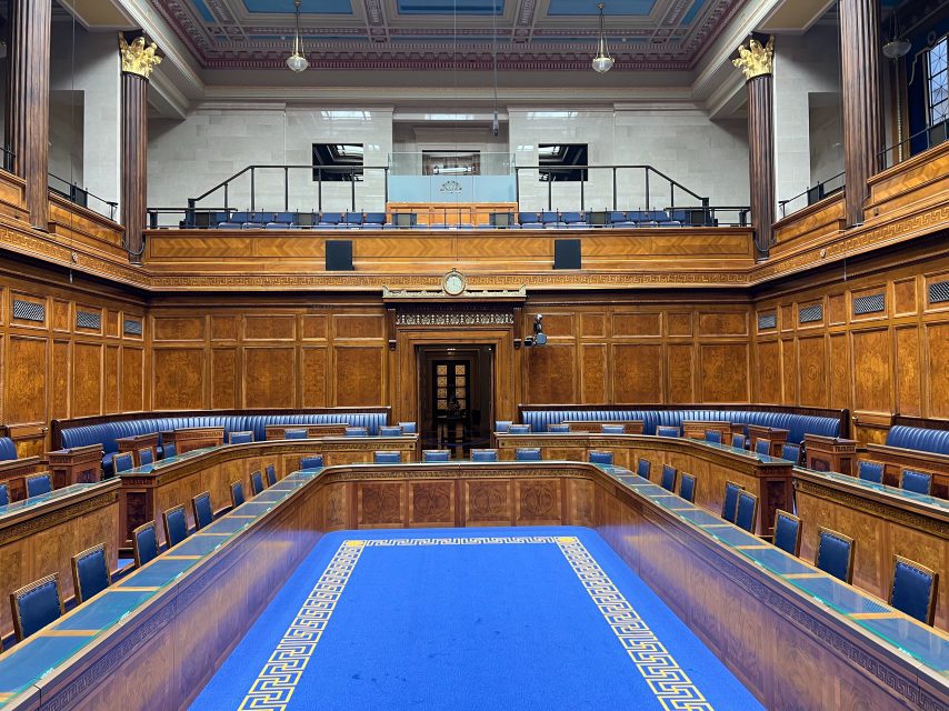 The debating chamber of the Northern Ireland Assembly, with wood panelling and tiered blue seating.