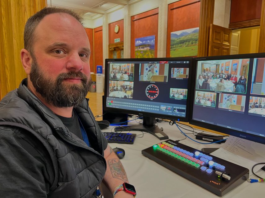 A technician at a broadcast control desk with dual monitors showing multi-camera feeds.