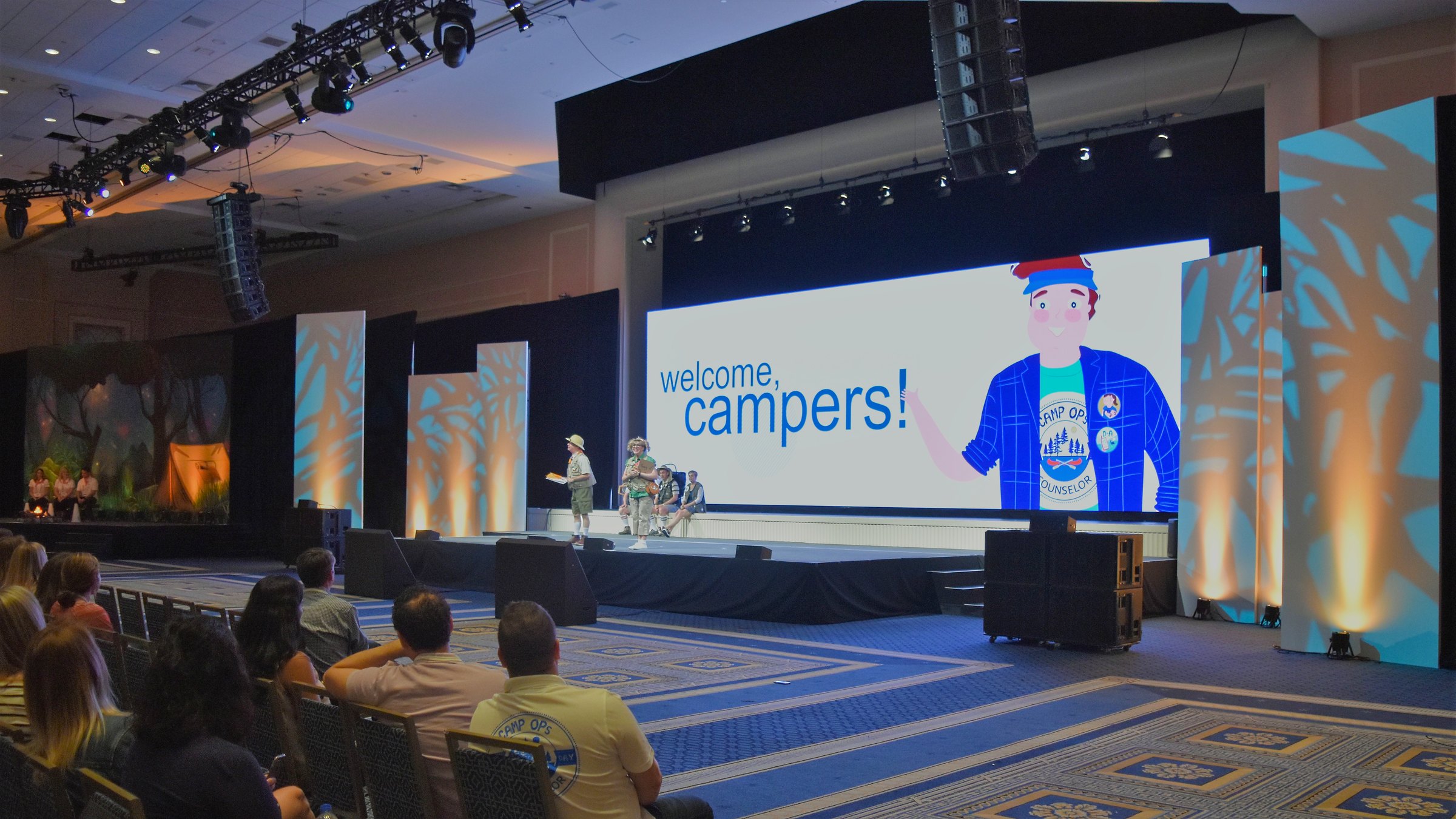 A hotel ballroom event stage with a large video wall reading 'welcome, campers!' beside rows of seated conference attendees.
