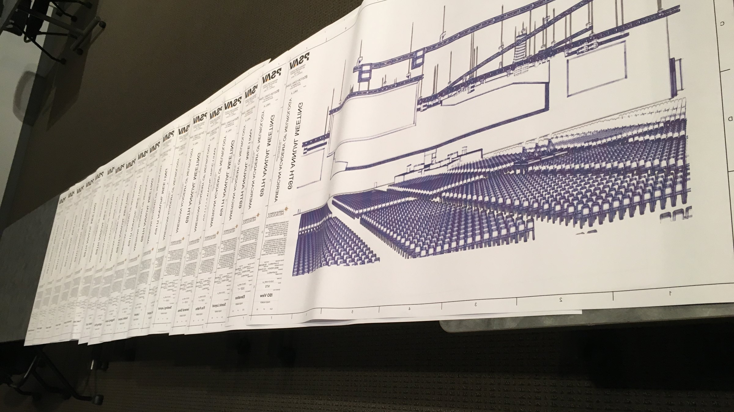 Large-format technical drawings spread on a table, with an isometric seating and stage layout in the foreground and a stack of title blocks behind.
